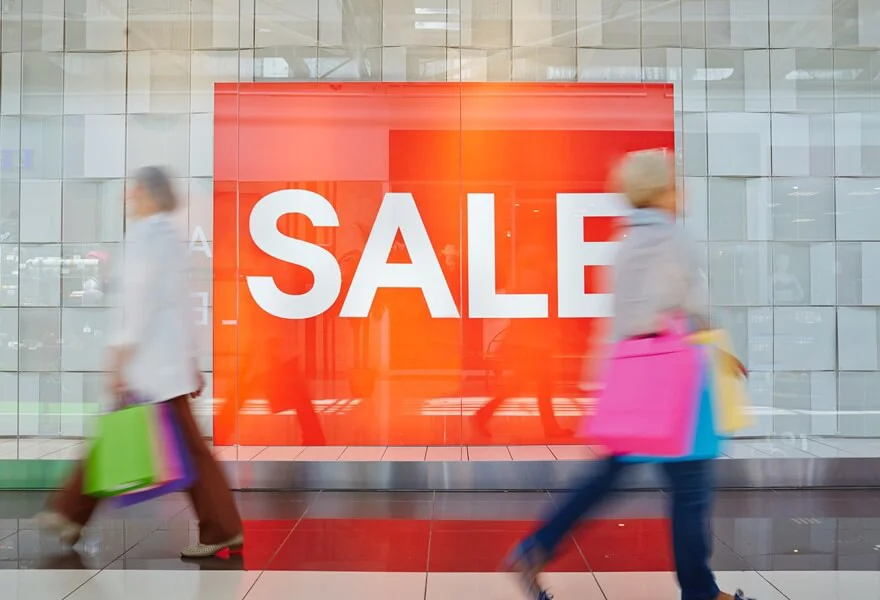 An image of shoppers walking past a big red sign with "SALE" written in big white letters.